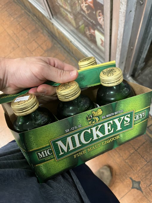 A hand holds a six-pack of Mickey's Fine Malt Liquor bottles featuring a green Mickey's branded carrier box.