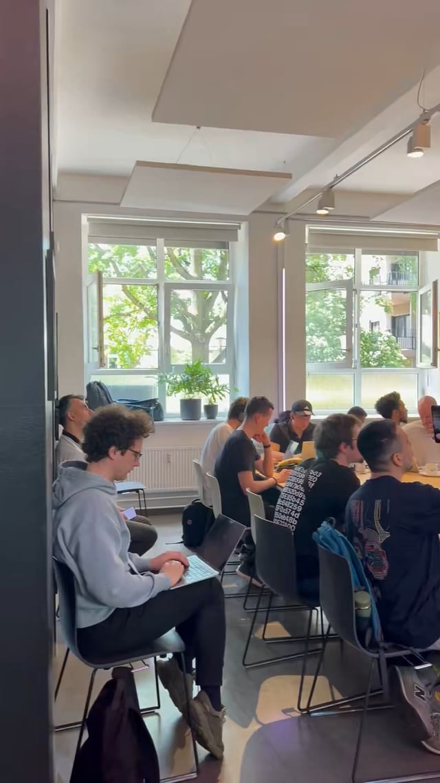 The image shows a group of people sitting around tables in a conference room with large windows that let in natural light. The attendees are engaged in a meeting or workshop focused on an Agentic payments hackathon, as indicated by the text "Algorand filling the room for their Agentic Payments Hackathon!". There is also a potted plant visible near one of the windows, adding some greenery to the scene.