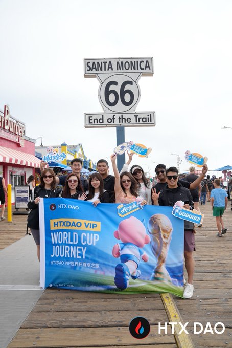 A group of HTX DAO VIP World Cup Journey travelers pose with celebratory signs under the iconic "Santa Monica 66 - End of the Trail" sign, showcasing the real-world rewards and community experiences that come with being an HTX DAO user.