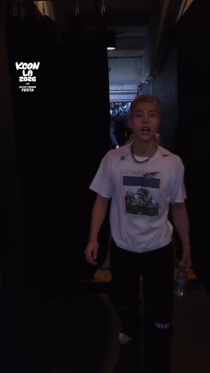 A man with pink/blonde hair wearing a graphic white t-shirt and silver chain walks backstage at KCON LA 2026, as indicated by the "KCON LA 2026 with Olive Young FESTA" logo overlaid on the dimly lit corridor. The image captures a candid Check Out Cam moment from Day 1 of the M COUNTDOWN event, showing an artist or performer leaving the venue.