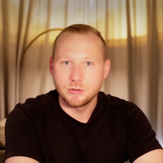 A close-up portrait of a fair-skinned man with short blonde hair and stubble, wearing a black t-shirt, with curtains visible behind.