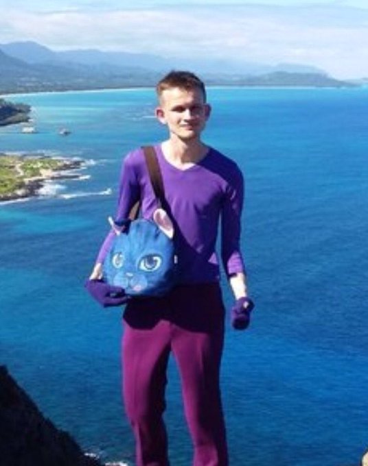 Photo of a young man in a purple top and pants standing against a bright blue sea backdrop, carrying a cat-shaped bag and wearing matching purple gloves.