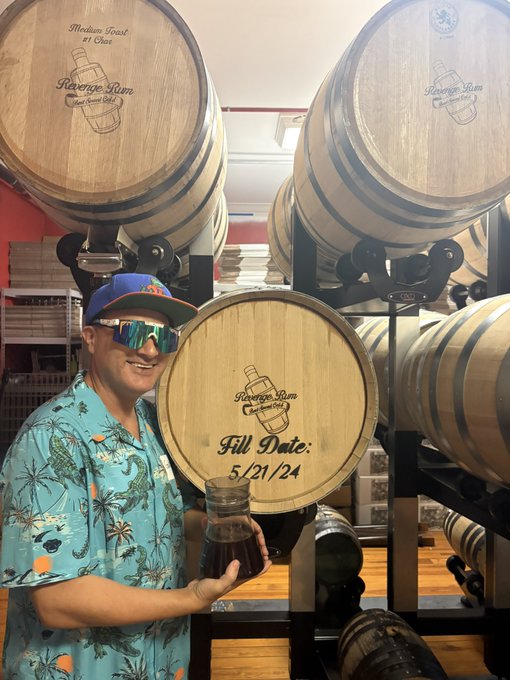 A man in a tropical shirt, sunglasses, and Mets cap stands in front of large wooden barrels labeled 'Riverside Rum Distillery,' holding a jar of dark liquid next to a barrel marked 'Fill Date: 5/21/24'.