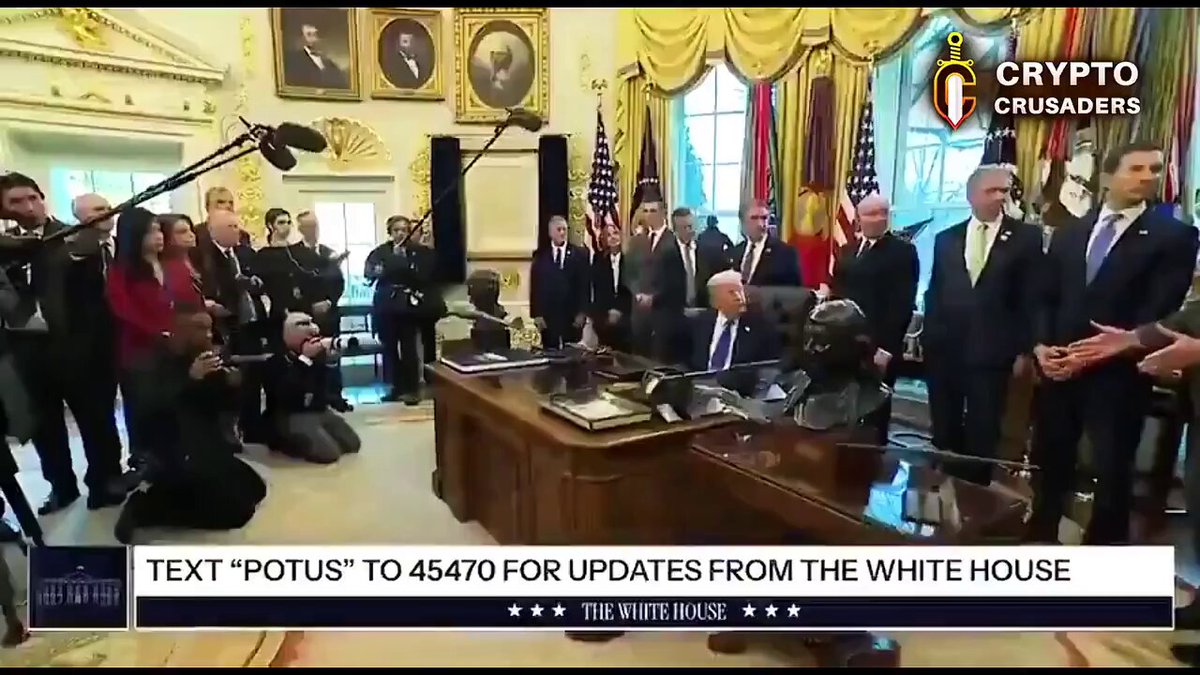 The image shows a group of people gathered around a desk, with some sitting down while others stand up. They appear to be engaged in a meeting or discussion related to crypto currency updates from the White House. The text "Text 'potus' to 4470 for updates from the white house" is visible on the image, indicating that they are discussing changes made by President Trump regarding the crypto market structure.