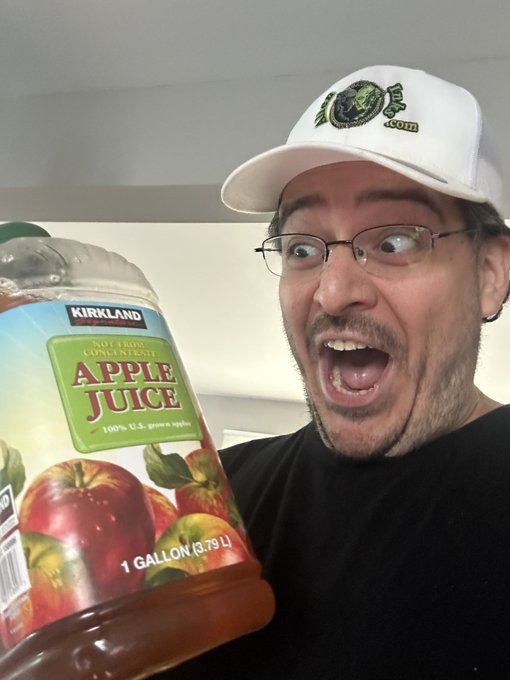A man with glasses and a white cap makes an exaggerated surprised expression while holding a 1-gallon Kirkland Signature apple juice bottle.