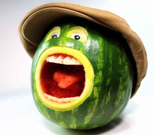 A carved watermelon designed to look like a cartoonish monster with wide eyes, a large mouth full of teeth, and a tongue, wearing a brown newsboy-style cap.
