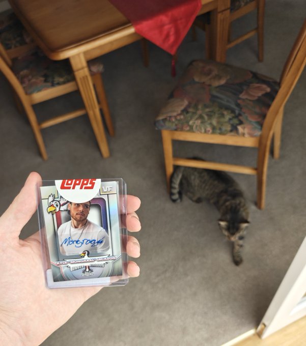 A hand holds up a Topps Nolan Ryan 2024 Chrome UFC card, with a tabby cat sitting on the carpet beneath a dining table and chairs in the background.