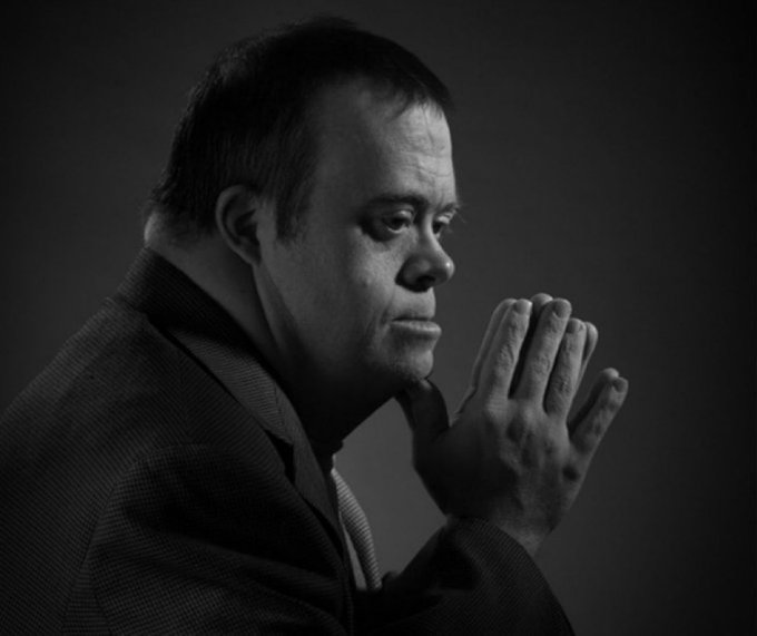 A black-and-white portrait photograph of a middle-aged man in profile, wearing a dark jacket, with his hands pressed together in a prayer-like gesture against a dark background.
