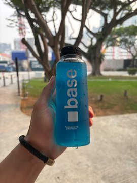 A hand holds a blue bottle labeled "base" in an outdoor park setting with trees and a body of water in the background.