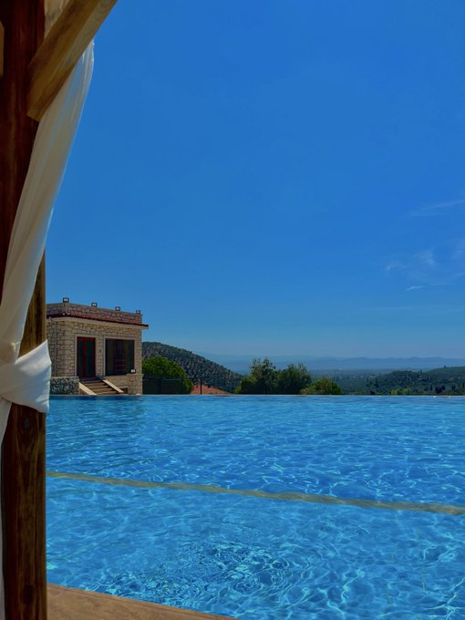 A scenic view of an infinity pool overlooking a vast landscape under a clear blue sky, with a stone building visible to the left.
