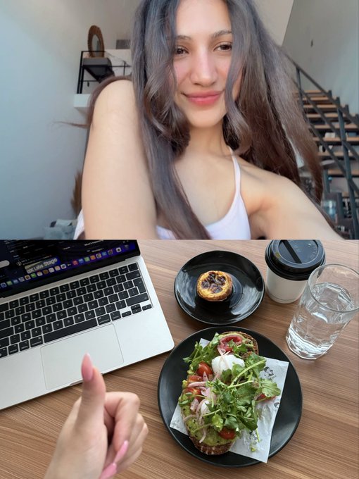 A selfie of a smiling young woman with long brown hair wearing a white tank top, with the lower half of the image showing a first-person view of a desk with a laptop, a salad on a black plate, a pastry, a coffee cup, and a glass of water.