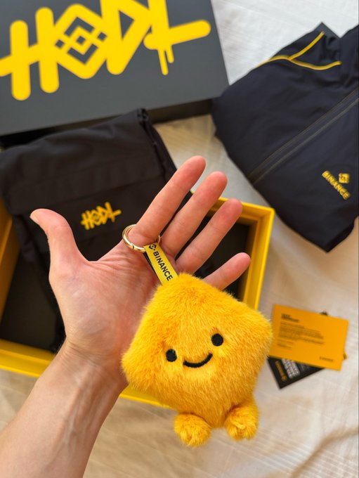 A hand holding a yellow fuzzy square-shaped Binance keychain charm with a smiley face, displayed against a yellow and black box and bag featuring 'HEX' branding.