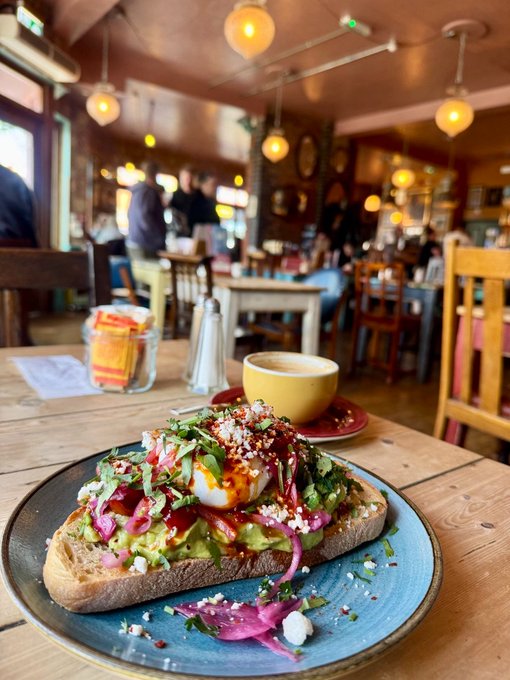 A vibrant avocado toast topped with poached eggs, pickled onions, feta cheese, and fresh herbs sits on a blue plate in a cozy café, accompanied by a yellow coffee cup on a wooden table.
