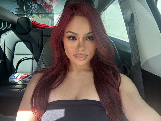 A selfie of a young woman with long red hair wearing a black strapless top, taken inside a car.