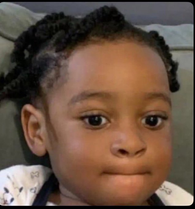 A close-up portrait of a young child with dark braided hair, wearing a white patterned shirt and looking slightly off-camera.