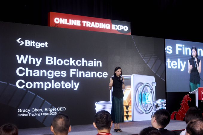 The image shows a woman standing at a podium, giving a speech or presentation about the benefits of blockchain technology for finance. The audience consists of several people sitting around her, attentively listening to her words. The setting appears to be an indoor event space with a stage and a large screen behind the speaker.