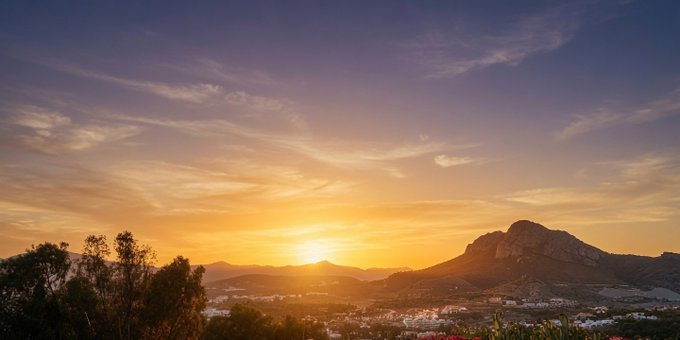 A golden sunset casts a warm glow over a mountainous landscape with a small town nestled in the valley below, silhouetted trees framing the left side of the scene. The dramatic sky and serene vista evoke the peaceful start of a new day, fitting the optimistic tone of "GM" (Good Morning).