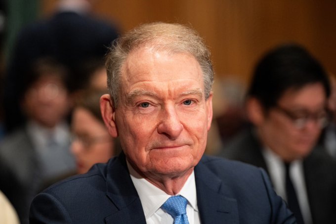 A close-up portrait of an older man with gray hair wearing a dark suit and blue tie, looking directly at the camera in what appears to be a formal hearing setting.