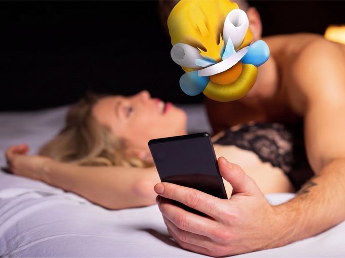 A shirtless man lies on a bed scrolling on a black smartphone while a woman in black lace lingerie lies behind him, with an emoji obscuring the man's face.
