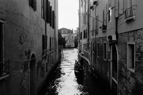 A black and white photograph of a narrow canal in Venice, flanked by tall buildings with weathered walls and shuttered windows.