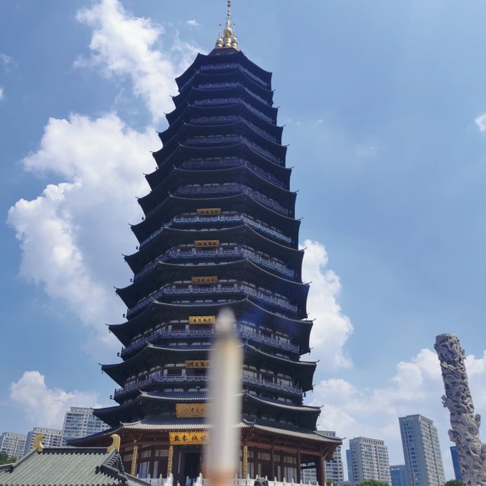 A tall, multi-tiered Chinese pagoda against a blue sky with clouds, featuring traditional architecture and an ornate stone pillar in the foreground.
