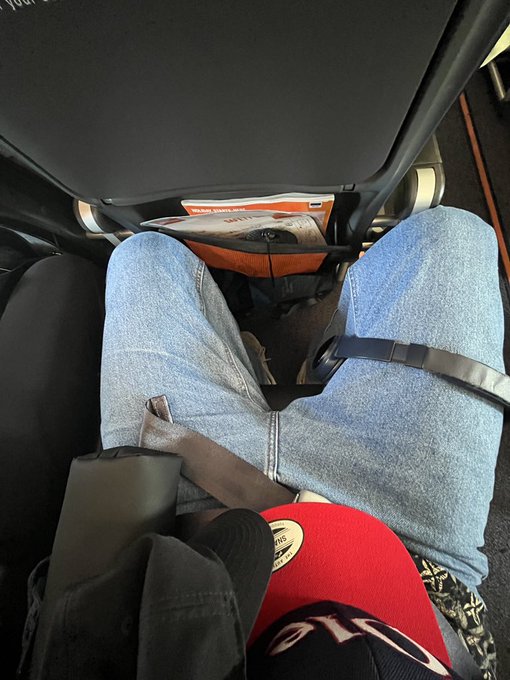 The image shows a person sitting in an airplane seat, wearing blue jeans and a red hat. There are two bags visible in the scene - one near the person's feet and another placed next to their seat. The legroom appears limited due to the presence of these bags, suggesting that the seating arrangement might not be ideal for long flights or extended periods of sitting.

The post "legroom" seems to be referring to the available space in front of the person seated on the airplane. It could also possibly indicate a preference for having more legroom during the flight, as some passengers may find it uncomfortable when there is limited space between their legs and the seat.