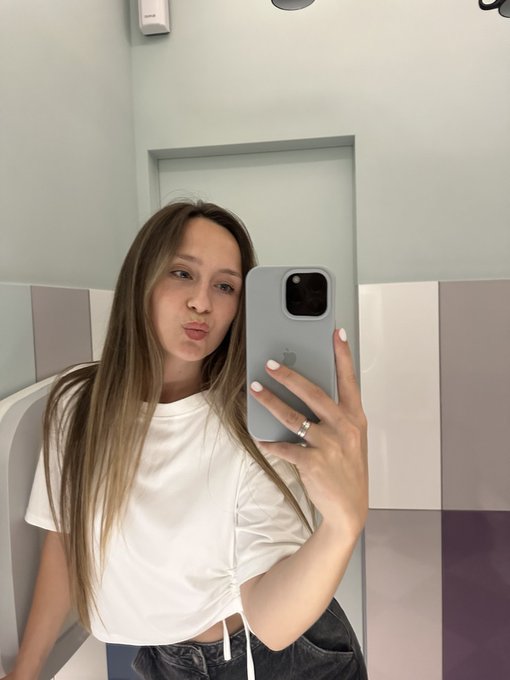 A mirror selfie of a young woman with long blonde hair wearing a white t-shirt and dark jeans, holding a smartphone in a bathroom with pastel tiled walls.