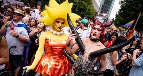 Two elaborately costumed individuals, one in an orange dress with a sun-ray headdress and another in demon attire with horns, posing at what appears to be a pride parade.