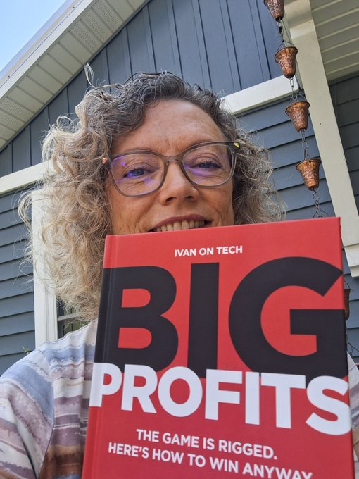 An outdoor selfie of an older woman with curly hair and glasses smiling behind a red book titled 'BIG PROFITS' by IVAN ON TECH with the subtitle 'THE GAME IS RIGGED. HERE'S HOW TO WIN ANYWAY'.