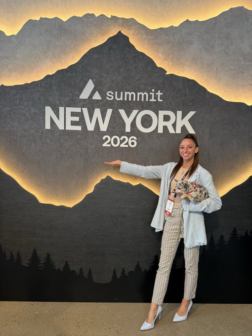 A smiling woman poses at the "Summit NEW YORK 2026" event backdrop featuring a mountain design, wearing a light blue blazer, plaid pants, and holding a small dog in a leopard print outfit.
