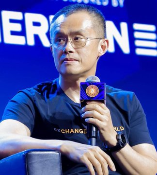 A man with glasses wearing a "CHANGE THE WORLD" t-shirt speaks into a Binance-branded microphone at what appears to be a crypto/blockchain event, aligning with the post's message about CZ (Changpeng Zhao, Binance's founder) expressing bullish sentiment on Bitcoin dips as buying opportunities.