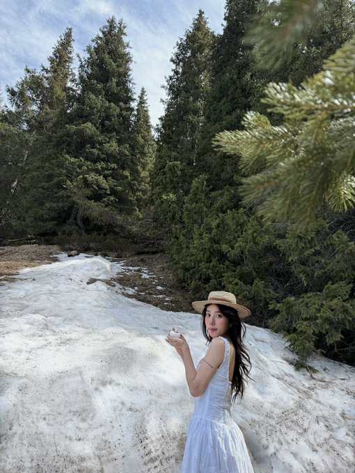 A young woman in a white sundress and straw hat stands on a snow patch in a forest, holding a snowball and smiling at the camera.