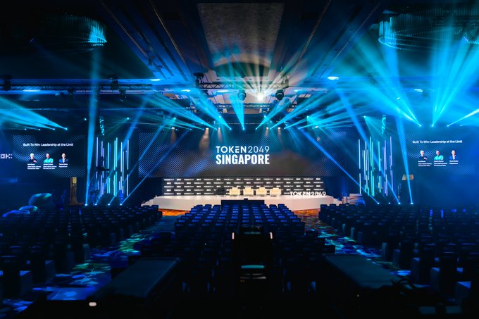 A grand conference hall is bathed in dramatic blue stage lights, with the main stage displaying "TOKEN2049 SINGAPORE" on the central screen flanked by side panels reading "Built To Win: Leadership at the Limit." The empty rows of seats and ready-to-go lighting setup correspond to the post's excitement about the OKX Main Stage, which will soon host notable speakers like Balajis, CryptoHayes, and Eric Trump when doors open tomorrow.