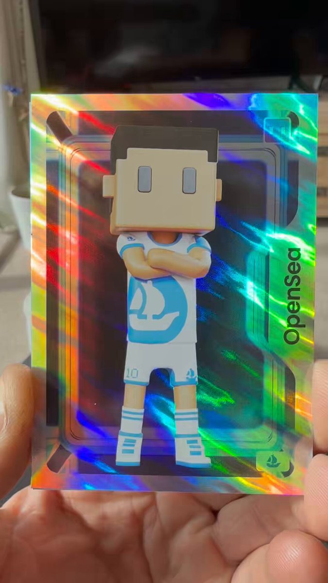 The image features a person holding up a small figurine of a soccer player, which appears to be made out of cardboard or foam board with a shiny surface that reflects light. The figurine has a white jersey and blue shorts, giving it an authentic appearance as if it were a real soccer player. The background is blurred, drawing attention to the figurine in focus. The post includes text such as "OpenSea" and "MeebitsNFTs", which may be related to the figurine or its creator.