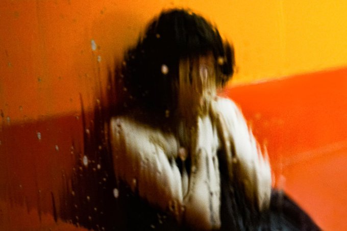 A painterly, blurred artwork of a figure in a white shirt against a vibrant orange and red background.