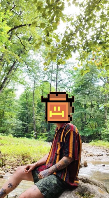 Person sitting by a rocky stream in a lush green forest, wearing a striped shirt and shorts, with their face obscured by a pixelated emoji sticker.