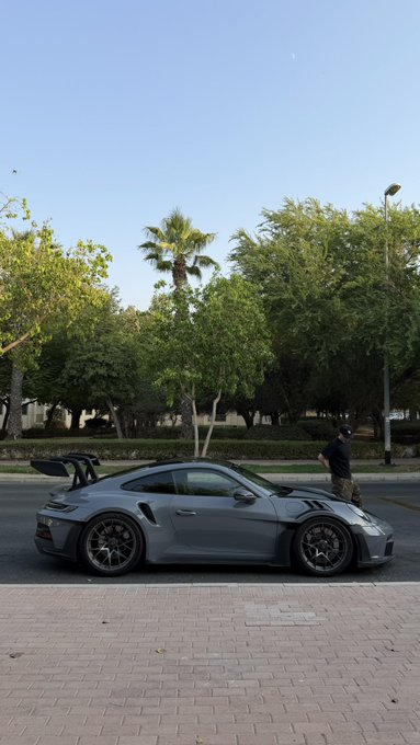 A gray Porsche 911 GT3 parked on a brick-paved area with a person standing nearby, trees and a street lamp in the background.