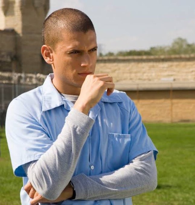 A young man with a buzz cut stands thoughtfully with his hand near his chin, wearing a light blue prison-style shirt over a gray long-sleeve shirt, set against an outdoor prison yard backdrop. The contemplative pose and setting humorously reflect the post's hesitation about swapping an INJ portfolio for ANSEM, as if weighing a risky decision that could land him behind bars.