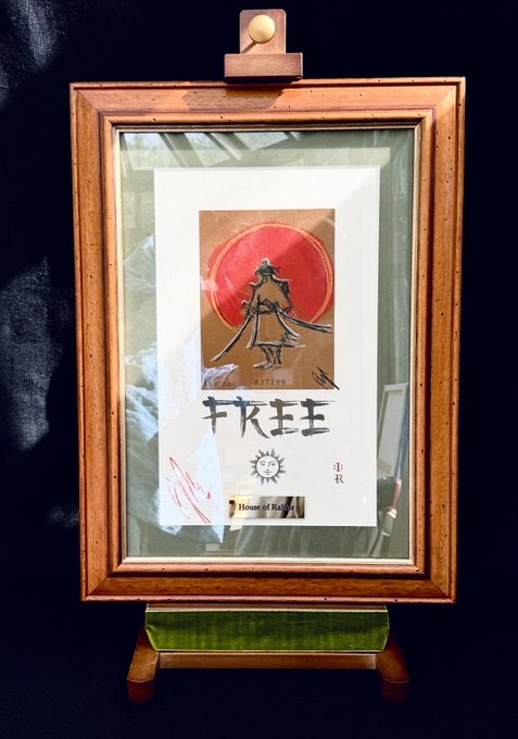 The image features a wooden frame with a painting of a person holding a sword, which appears to be a Samurai. The painting has a red background and white text that reads "FREE Samurai". The frame is placed against a black background, making the colors of the painting stand out even more. There are also some words written in another language on the bottom right corner of the image.