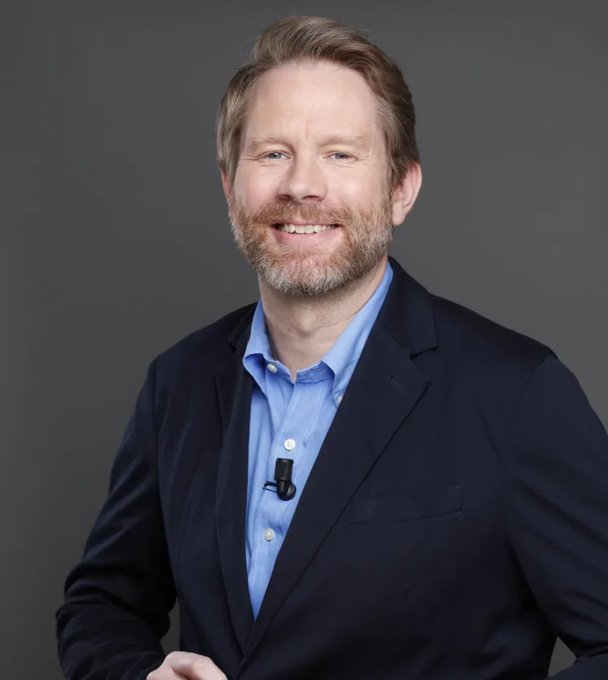 The image shows a smiling, bearded man in a dark navy blazer and light blue button-down shirt, with a small lapel microphone clipped to his shirt, set against a plain gray background—likely a professional headshot of Eric Larch, the founder of The Bitcoin Society referenced in the post about the Coldcard hack and the safety of the company's BTC holdings.