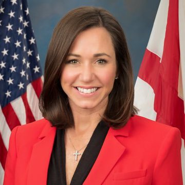 Senator Katie Britt, wearing a red blazer and cross necklace, smiles in front of the American flag and Alabama state flag as she advocates for the CLARITY Act to secure U.S. leadership in digital finance innovation.