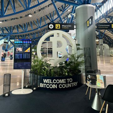 The image shows a large circular sign with the words "Welcome to Bitcoin Country" written on it, surrounded by potted plants. There are chairs placed around the area where people can sit and read about Bitcoin. The scene also includes other signs and displays related to Bitcoin in the background.