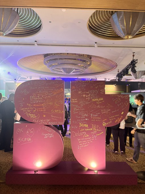 Two large pink heart-shaped signs covered in handwritten messages and names, displayed at an indoor event with people in the background.