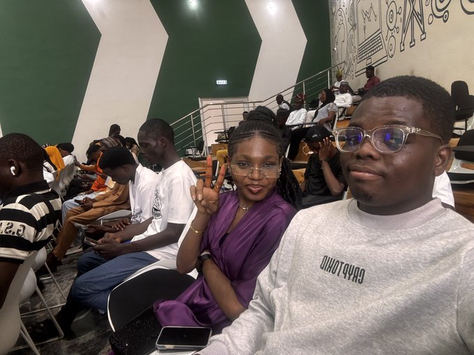 A young couple poses for a selfie in an auditorium, with the woman in a purple satin top flashing a peace sign and the man wearing glasses and a grey sweatshirt that reads 'JINOT4V4O'.