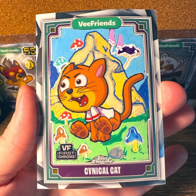 The image features a hand holding up a sticker of a cartoon cat with orange fur, wearing a white shirt and hat. The cat appears to be running or jumping around, surrounded by mushrooms and other elements typical of a forest scene. The background is blurred, but there are several other stickers visible in the image as well. The post reads "Mushroom Mountain Let's see those NFT scenes @veefriends @VeeFriendsCards".