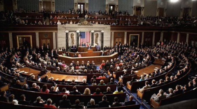 A wide interior view of the United States House of Representatives chamber, filled with members of Congress seated at their desks facing the Speaker's rostrum with the American flag.