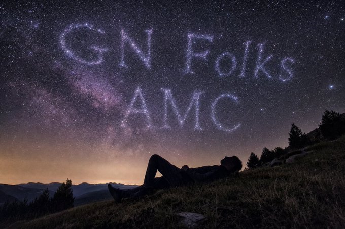 A person lies on a grassy hillside gazing up at a starry night sky where the Milky Way is visible and the words 'G N Folks AMC' appear formed by stars.