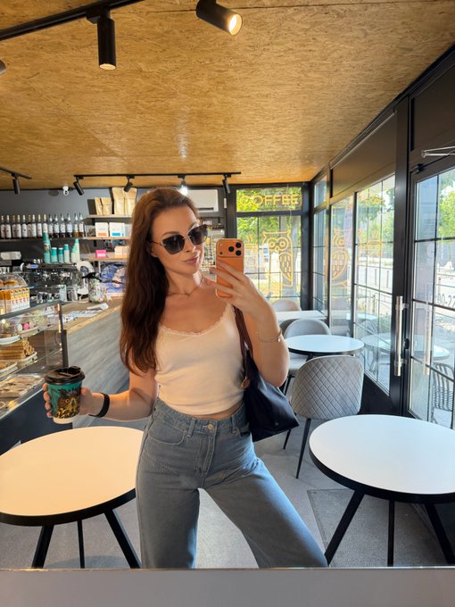 A young woman wearing sunglasses, a white tank top, and jeans taking a mirror selfie with an orange phone inside a coffee shop, while holding a colorful reusable cup.