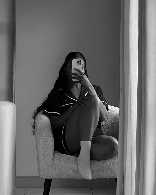 A black-and-white mirror selfie of a woman sitting curled up in a textured armchair, wearing a dark pajama set and white socks while holding a smartphone to cover her face.