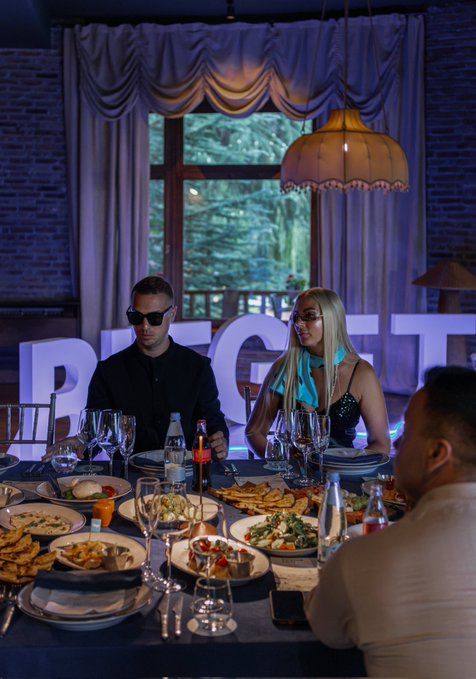 Evening dinner scene at a Bitget branded event, with a man in sunglasses and a blonde woman seated at a candlelit table set with wine glasses and assorted dishes in a venue with draped curtains and a glowing pendant lamp.